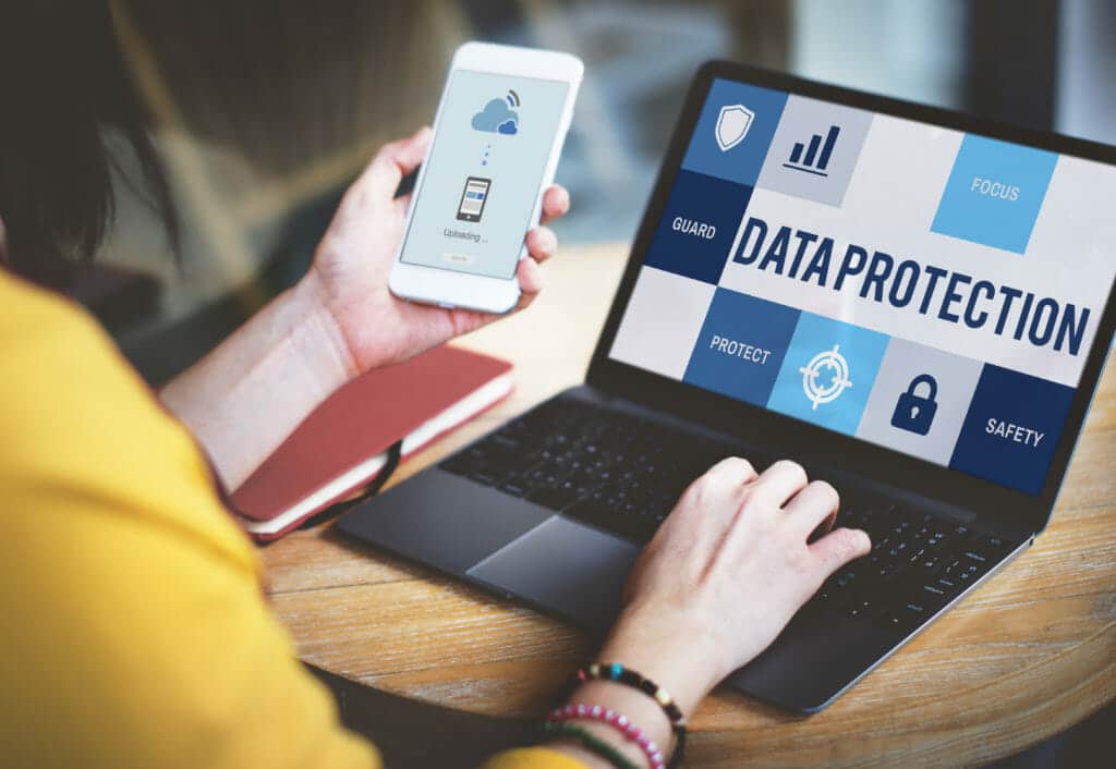 woman using a laptop and a phone at the same time, the laptop has the text 'data protection' on it and the phone has a phone image connecting to the cloud