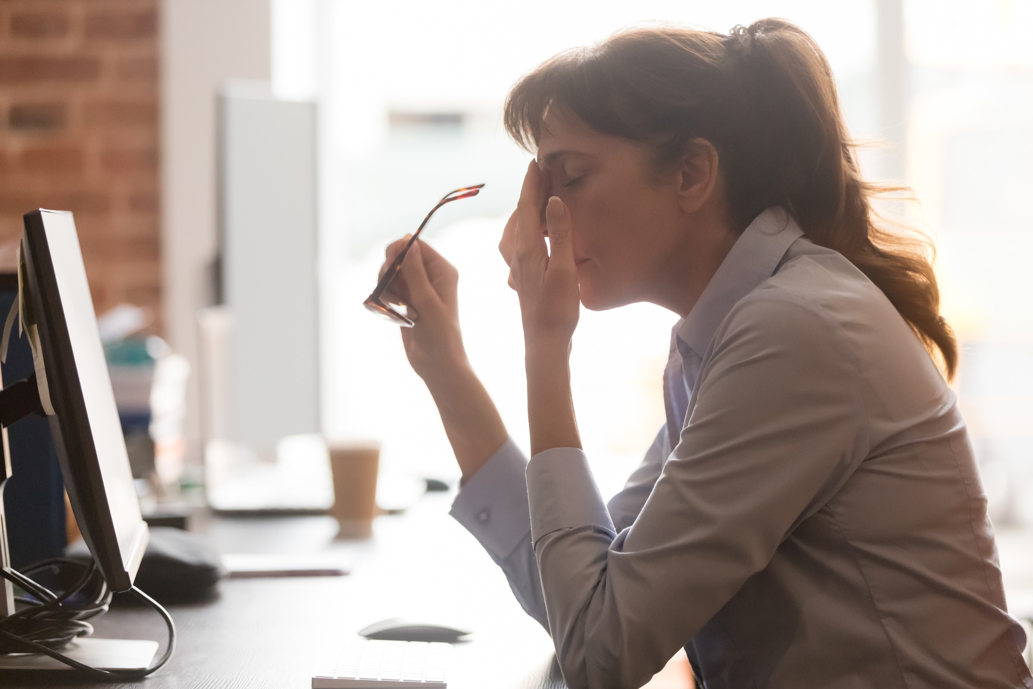 Exhausted female worker sit at office desk take off glasses feel unwell having dizziness or blurry vision, tired woman employee suffer from migraine or headache unable to work. Health problem concept