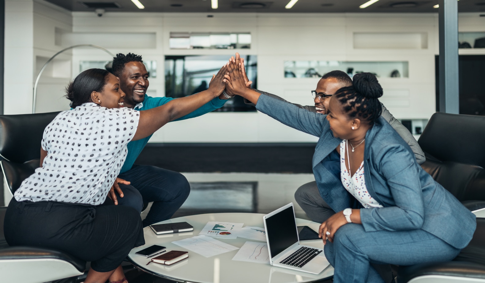 four people giving eachother a highfive whilst sat in an office working