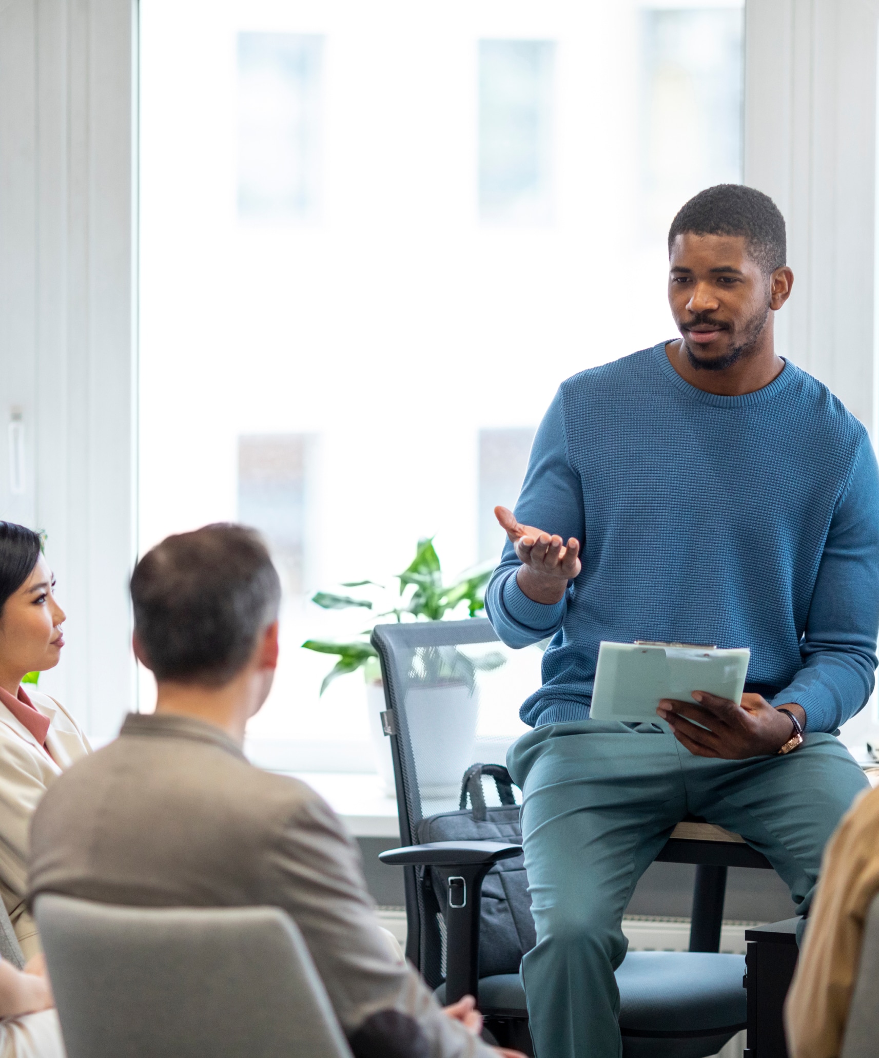 Man sat on the edge of a desk holding a clip board and speaking to his colleagues