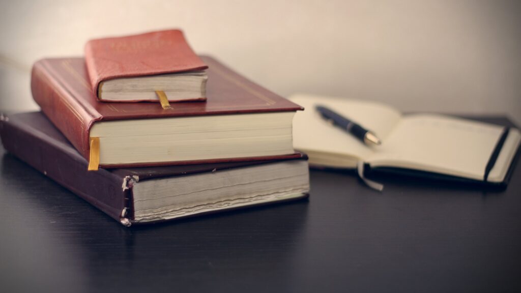 Pile of three leather bound books in the foreground with one open book and a pen in the background