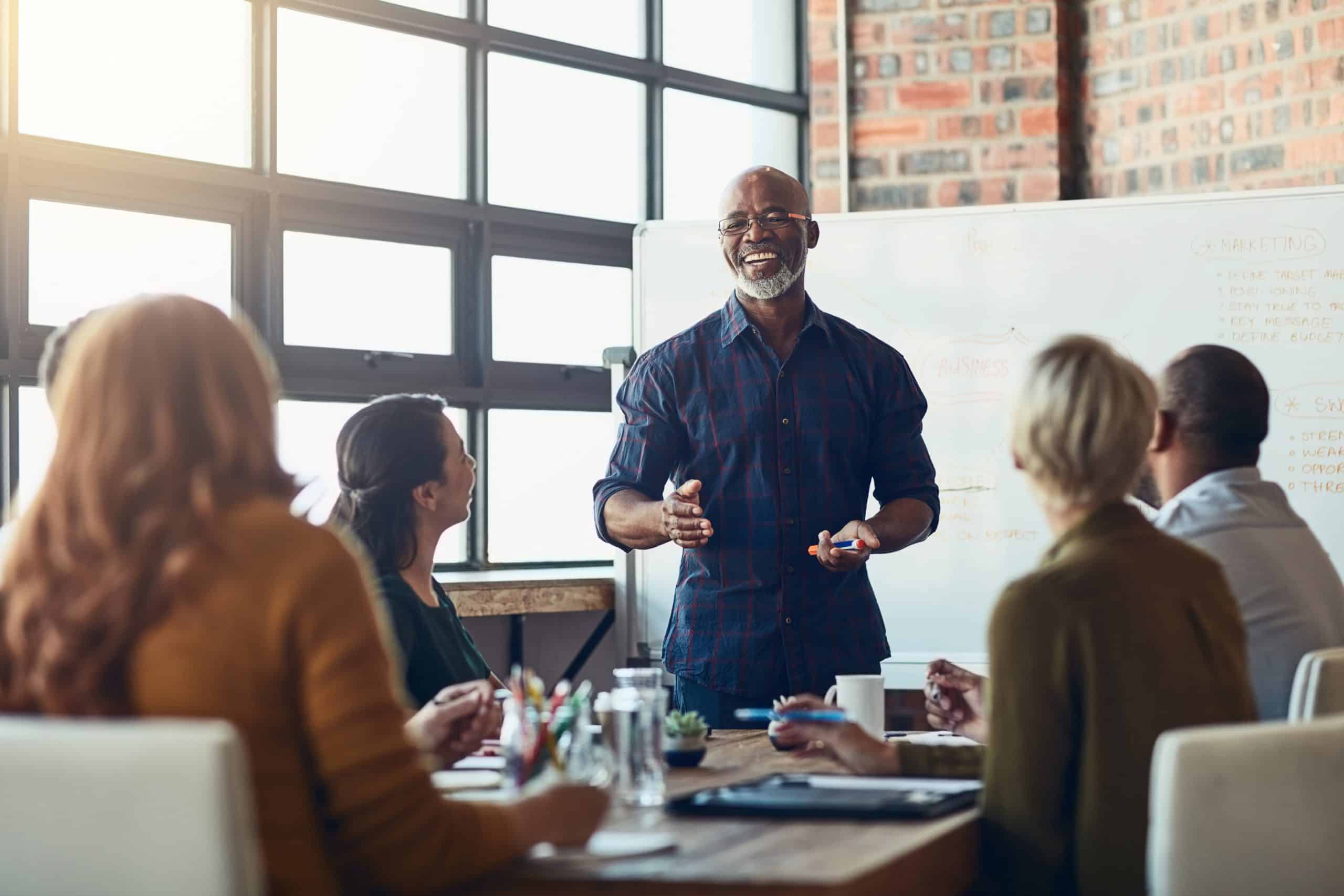 Smiling man leading a presentation to a group of people in an office in front of a white board