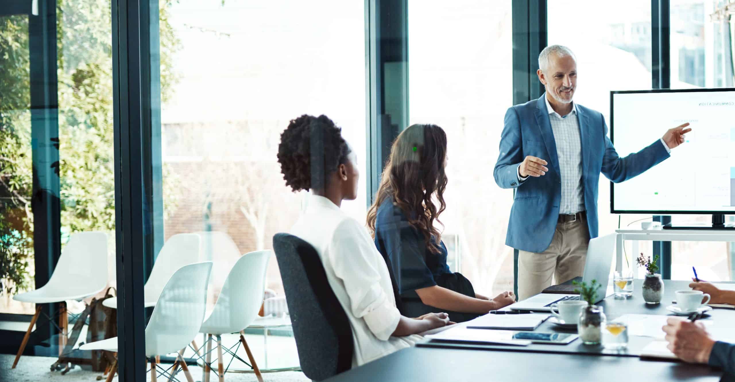 Man standing at the head of a table leading a presentation in the office to his colleagues who are sat around the table