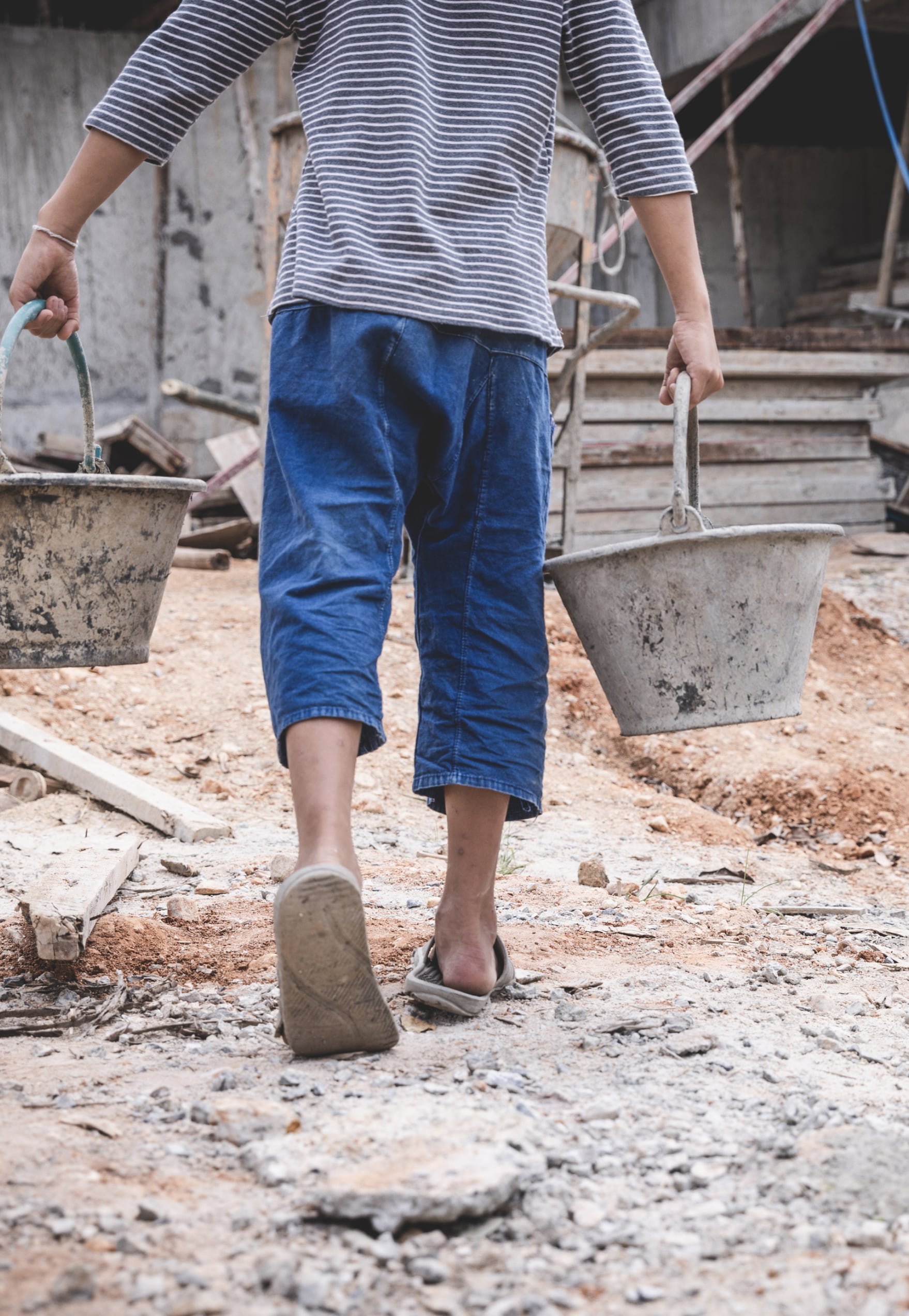 The back of a child carrying two buckets of water across a a desolate area