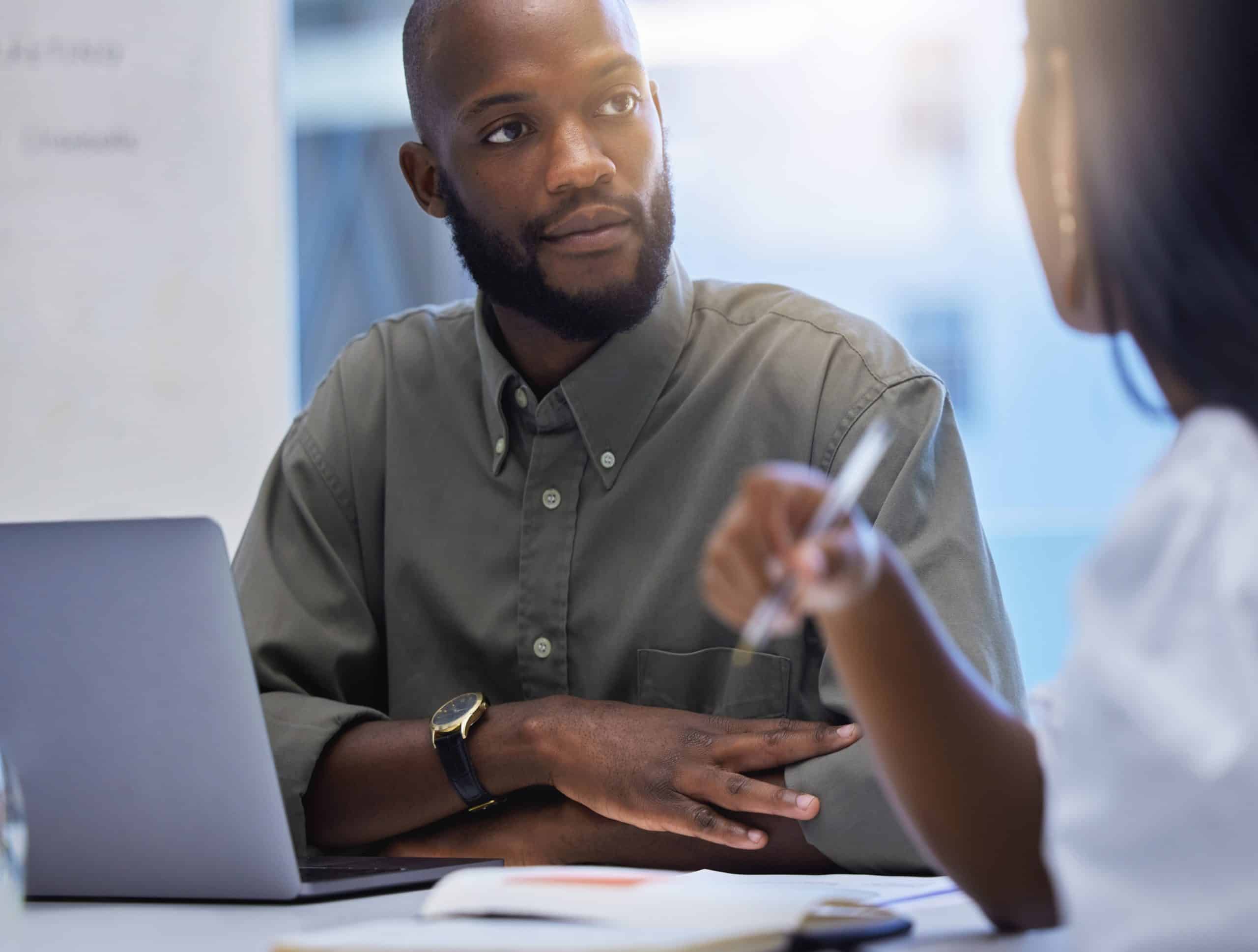 Serious looking man sitting at a desk being spoken to by a women opposite him